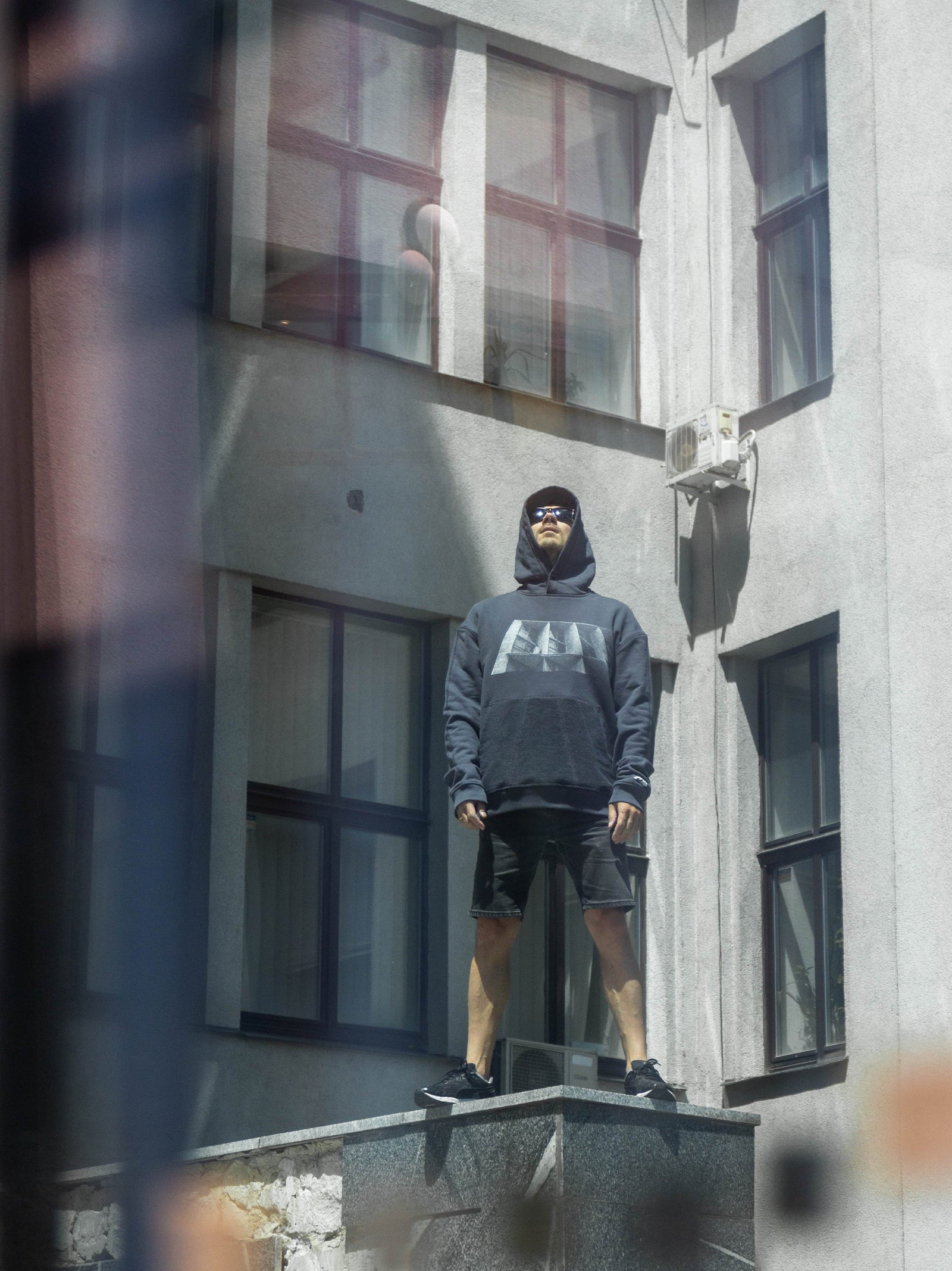 Person wearing a dark hoodie with a brutalist print in front of a building.