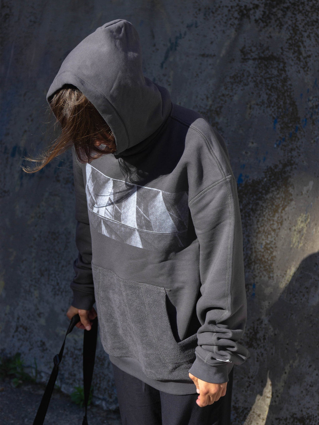 Person wearing a gray hoodie with a brutalist print against a textured wall.