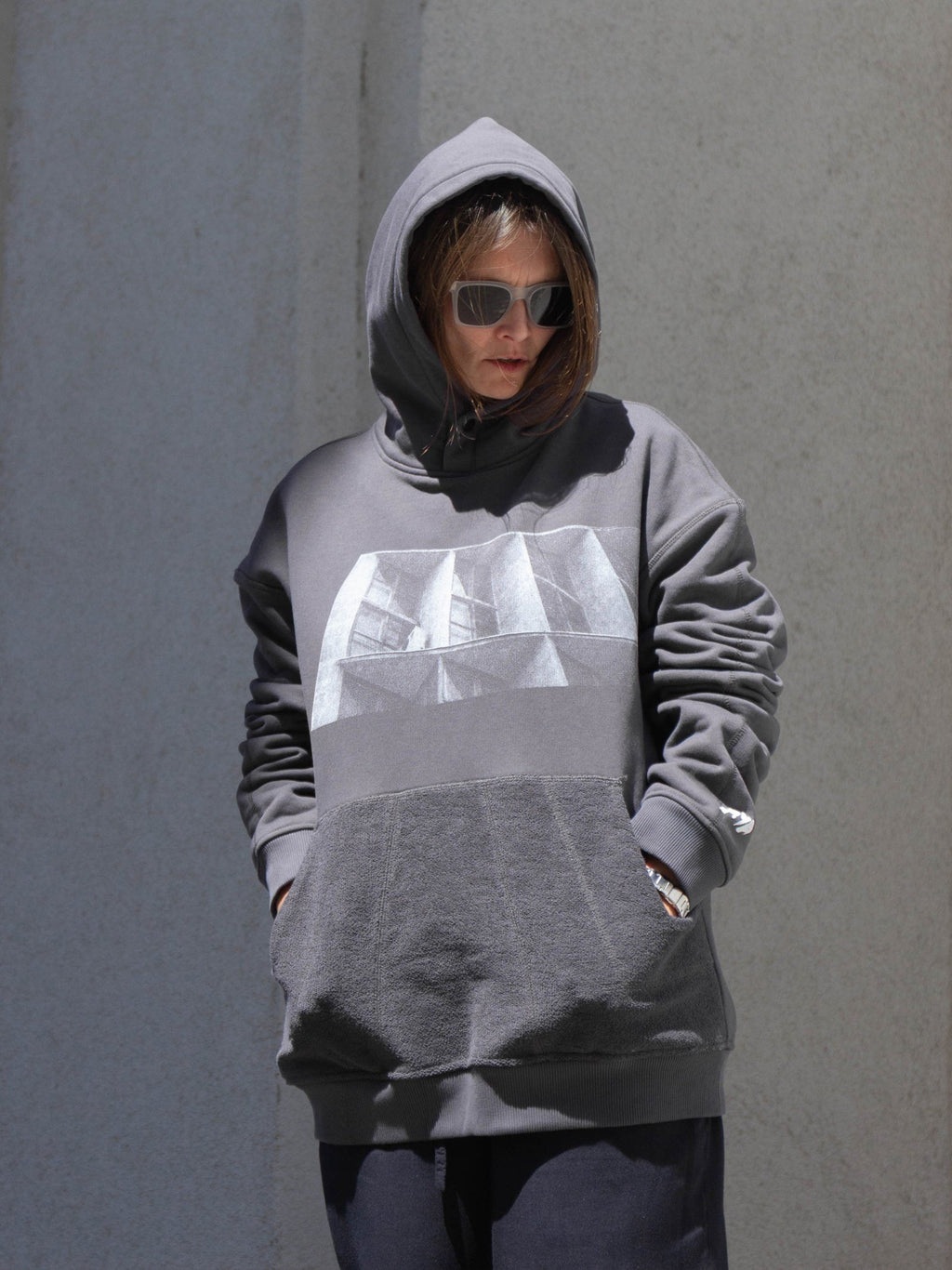 A person wearing a gray oversized hoodie with a large white geometric graphic on the chest, standing against a textured gray wall.