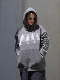 A person wearing a gray oversized hoodie with a large white geometric graphic on the chest, standing against a textured gray wall.