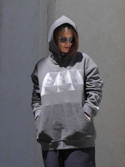 A person wearing a gray oversized hoodie with a large white geometric graphic on the chest, standing against a textured gray wall.